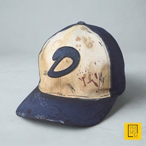 May include: A navy blue baseball cap with a curved brim. The cap features a tan front panel with a dark blue letter "D" and distressed-style graphics. The cap's design includes a dark blue brim and a button on top.