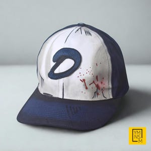 May include: A navy blue and white baseball cap with a dark blue letter "D" and red splatter details on the front panel. The cap has a classic six-panel design and a navy blue brim.