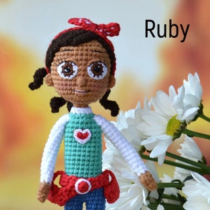 May include: Two crocheted dolls with brown skin and black hair. The doll on the left is wearing a blue shirt with a red heart, blue jeans, and a red belt. The doll on the right is wearing a white shirt, a green shirt with a red heart, blue jeans, a red belt, and a red headband. The dolls are standing in front of a bouquet of white daisies.