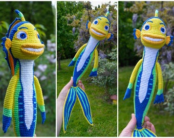May include: Crocheted fish toy in yellow, blue, and green. The fish has a smiling face, fins, and a tail. The toy is handmade and appears to be soft and cuddly. The background is a blurred outdoor scene.