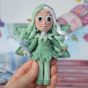 Può includere: Una bambola di fata di uncinetto verde con capelli bianchi, vestita con un vestito verde e che tiene una bacchetta blu.