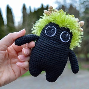 Puede incluir: Un juguete de peluche negro tejido a crochet con un peinado peludo verde y marrón. El juguete tiene grandes ojos negros y una boca sonriente blanca.