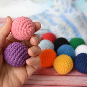 May include: A hand holds two crocheted balls, one pink and one purple. Several other colorful crocheted balls in red, orange, black, blue, green, white, and yellow are in the background. The balls are likely for a craft project or a child's toy.