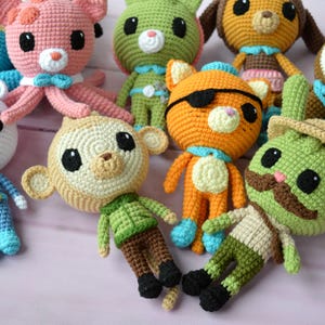 Puede incluir: Una colección de ocho animales de peluche tejidos a crochet, que incluyen un pulpo rosa, un mono verde, un perro marrón, un conejo verde, un oso polar blanco, un extraterrestre verde, un gato naranja y un panda marrón. Los animales llevan todos atuendos diferentes y tienen diferentes expresiones.