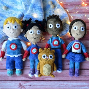 May include: Five crocheted plush toys, including four human figures and a cat. The human figures are wearing blue and red outfits with a circular emblem on the chest. The cat is orange and white. The toys are arranged on a wooden surface.