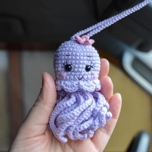 May include: A purple crocheted octopus with a pink flower on its head. It has black eyes and a smiling mouth. The octopus is hanging from a string.