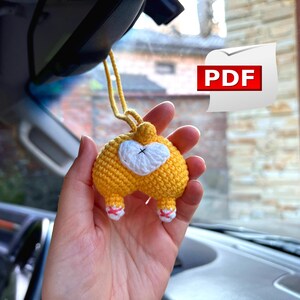 May include: A crocheted yellow dog rear end with white tail and pink feet hanging from a rearview mirror in a car. The dog is a car charm.