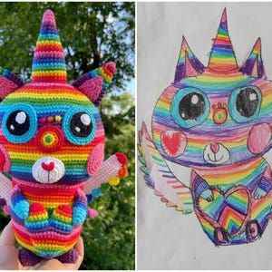 May include: A rainbow crochet cat with wings and a unicorn horn. The cat is holding a heart in its paws. The cat is next to a drawing of a similar cat on a white t-shirt.