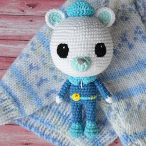 May include: A white crocheted stuffed animal with blue and green accents. The animal is wearing a blue jumpsuit with a yellow star on the chest. The animal is sitting on a blue and white knitted blanket.