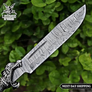 May include: A Damascus steel knife with a detailed blade pattern and a handle featuring a wolf's head design. The knife has a long, sharp blade and is set against a backdrop of green foliage. The text "Legends of Vikings" is visible.