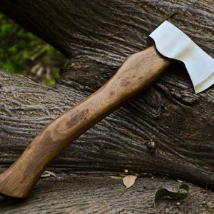 May include: A handcrafted axe with a polished steel blade and a wooden handle. The axe is resting on a weathered, textured tree trunk. The handle is a warm brown color, and the blade is silver.