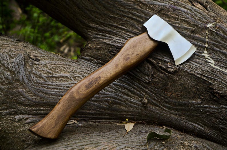 May include: A handcrafted axe with a polished steel blade and a wooden handle. The axe is resting on a weathered, textured tree trunk. The handle is a warm brown color, and the blade is silver.