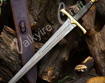 May include: A long, ornate sword with a patterned blade, gold-colored crossguard and pommel, and a black grip. It rests beside a brown leather sheath with decorative stitching. The sword is displayed against a backdrop of wooden logs.