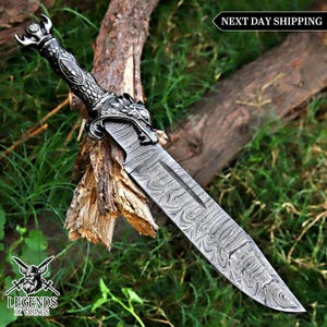 May include: A decorative dagger with a patterned blade and a dragon-themed handle. The handle is silver and black, and the blade has a unique swirling pattern. The dagger is displayed on a piece of wood with green foliage in the background.