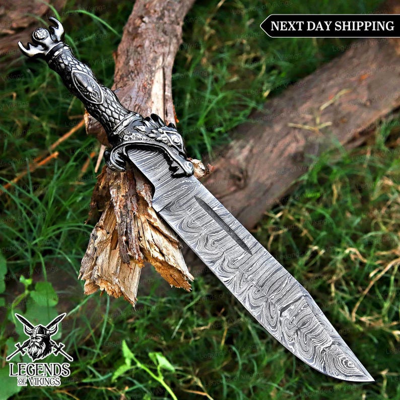 May include: A decorative dagger with a patterned blade and a dragon-themed handle. The handle is silver and black, and the blade has a unique swirling pattern. The dagger is displayed on a piece of wood with green foliage in the background.