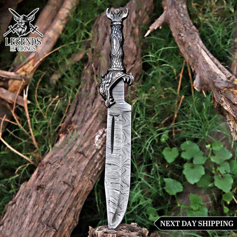 May include: A detailed, silver-colored Viking dagger with a patterned blade and ornate handle. The handle features a dragon head design. The dagger is displayed on a wooden surface with a green background. The text "Legends of Vikings" is in the upper left corner.