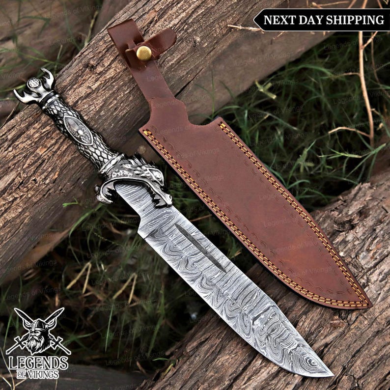 May include: A decorative dagger with a patterned blade and ornate silver handle featuring a dragon design. The dagger is paired with a brown leather sheath. The image includes the text "Legends of Vikings" and "Next Day Shipping".