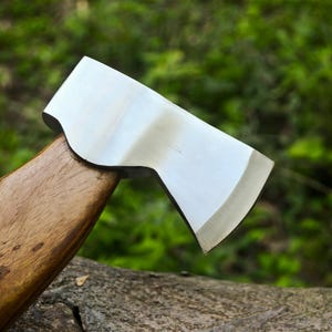 May include: Close-up of a hatchet with a polished steel blade and a wooden handle. The axe head is silver, and the handle is a warm brown color. The hatchet is resting on a log with a blurred green background.