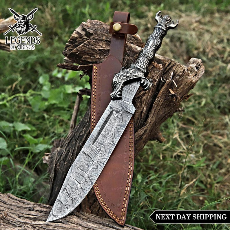May include: A decorative dagger with a patterned blade and ornate silver handle, resting in a brown leather sheath. The handle features a detailed design, and the blade has a unique swirling pattern. The sheath is secured with a leather strap.