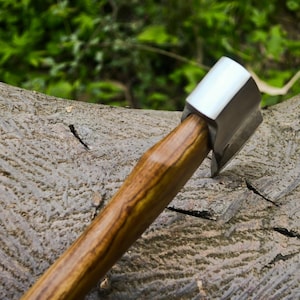 May include: A small axe with a polished metal head and a wooden handle. The handle has a natural wood grain pattern. The axe is resting on a textured, gray tree trunk, with green foliage in the background.