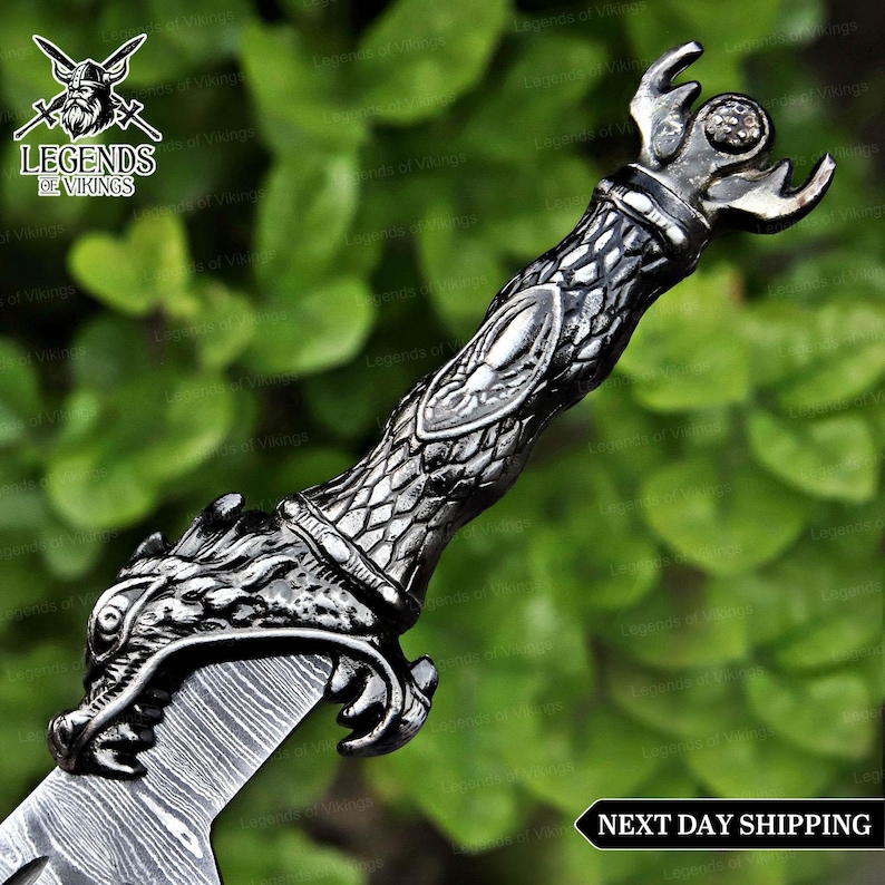 May include: Close-up of a decorative sword with a detailed, silver-colored handle. The handle features a dragon head design and intricate patterns. The blade has a textured appearance. The image is set against a blurred green background.