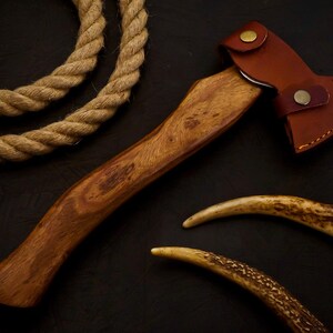 May include: A small, handcrafted axe with a wooden handle and a brown leather sheath. The axe head is metal. Two curved antler pieces and a length of rope are also present.