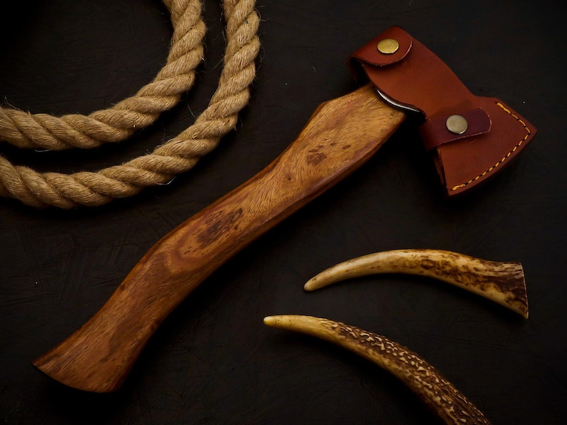 May include: A small, handcrafted axe with a wooden handle and a brown leather sheath. The axe head is metal. Two curved antler pieces and a length of rope are also present.