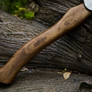 May include: A handcrafted axe with a wooden handle and a silver-colored blade. The handle is a warm brown color with visible wood grain. The axe is resting on a textured, dark-colored surface, possibly a tree trunk.
