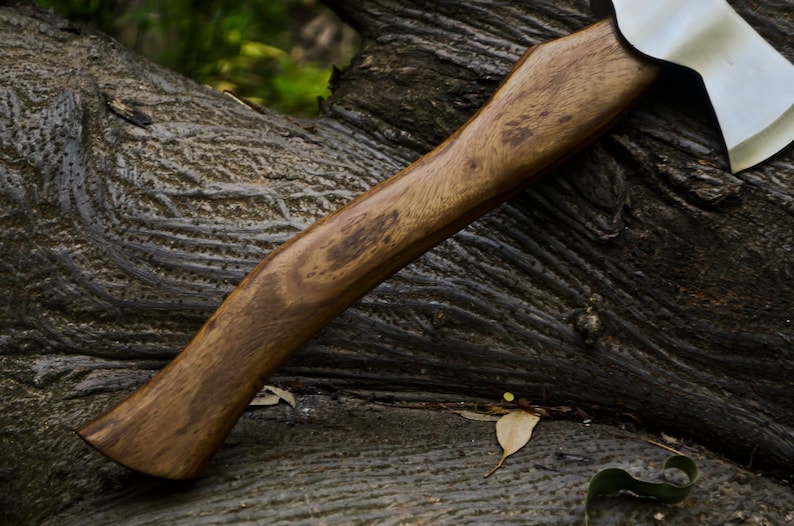 May include: A handcrafted axe with a wooden handle and a silver-colored blade. The handle is a warm brown color with visible wood grain. The axe is resting on a textured, dark-colored surface, possibly a tree trunk.