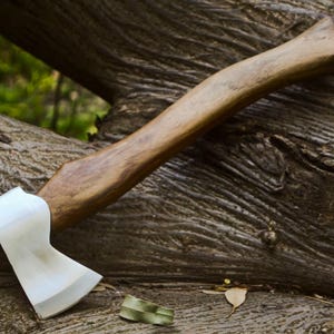 May include: A handcrafted axe with a polished steel blade and a wooden handle. The axe is resting on a weathered tree trunk, showcasing its sharp edge and sturdy construction. The handle is approximately 15 inches long.