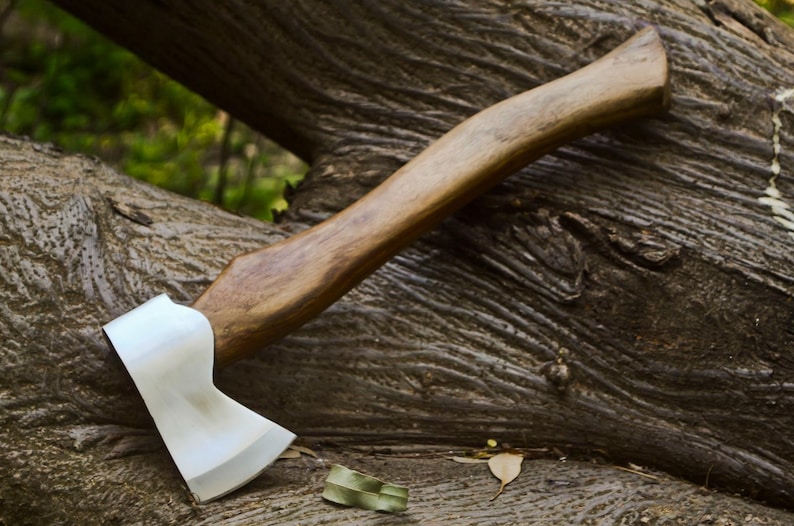 May include: A handcrafted axe with a polished steel blade and a wooden handle. The axe is resting on a weathered tree trunk, showcasing its sharp edge and sturdy construction. The handle is approximately 15 inches long.