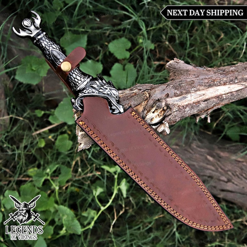 May include: A decorative knife with a silver handle featuring a dragon design and a brown leather sheath. The knife has a curved blade and is secured with a leather strap. The sheath is stitched with a decorative pattern. The image includes the text "Legends of Vikings".