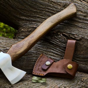 May include: A handcrafted axe with a wooden handle and a polished steel blade. The axe is accompanied by a brown leather sheath. The axe and sheath are resting on a textured wooden surface.