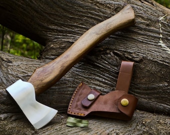 Puede incluir: Un hacha artesanal con mango de madera y hoja de acero pulido. El hacha va acompañada de una funda de cuero marrón. El hacha y la funda descansan sobre una superficie de madera texturizada.