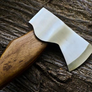May include: A close-up of a hatchet with a polished steel blade and a wooden handle. The hatchet is set against a textured, dark brown wooden surface, highlighting the tool's sharp edge and craftsmanship.