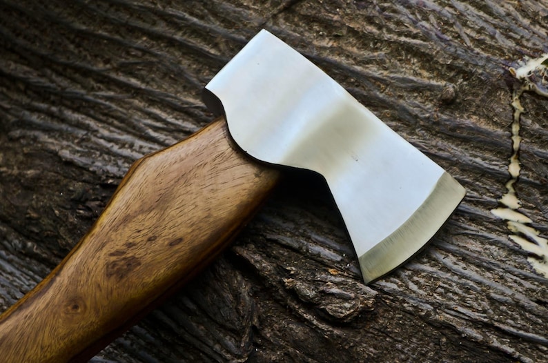 May include: A close-up of a hatchet with a polished steel blade and a wooden handle. The hatchet is set against a textured, dark brown wooden surface, highlighting the tool's sharp edge and craftsmanship.