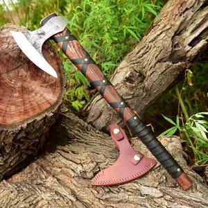 May include: A handcrafted axe with a wooden handle wrapped in black leather and accented with brass studs. The axe head is silver and has a curved blade. A brown leather sheath protects the blade. The axe rests on a tree trunk.