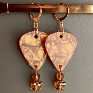 May include: A pair of gold-tone earrings with a guitar pick-shaped pendant and a dangling amber-colored bead. The guitar pick pendant has a marbled, copper-toned design.
