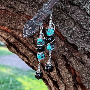 May include: Long dangle earrings with alternating black and turquoise-colored beads, suspended from a silver-toned wire. The earrings are displayed on a silver spoon against a blurred natural background. The earrings have a unique, handcrafted appearance.