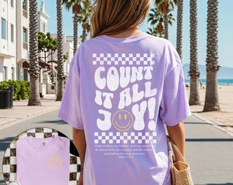 May include: A lilac-colored t-shirt with the words "COUNT IT ALL JOY!" in white, a yellow smiley face, and a checkered pattern. The shirt also features a small smiley face on the front. The text "Count it all joy..." is printed below the graphic.