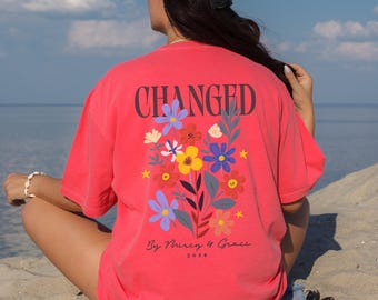 May include: A coral-colored t-shirt with the word "CHANGED" in black block letters. Below the text is a floral design with blue, yellow, and white flowers. The text "By Mercy & Grace 2023" is printed below the floral design.