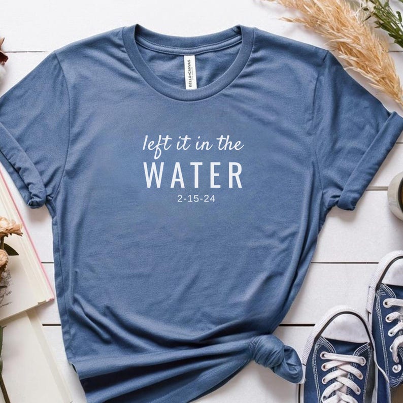 May include: A blue t-shirt with the text "left it in the WATER" and the date "2-15-24" printed in white. The shirt is made of a soft material and has a crew neck. The sleeves are rolled up. A pair of blue sneakers are visible in the bottom right corner.
