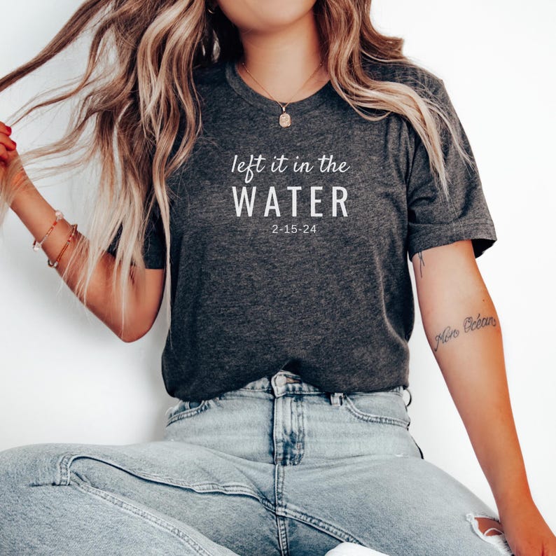 May include: Dark gray t-shirt with the text "left it in the WATER" and the date "2-15-24" in white. The shirt is paired with light blue jeans.