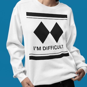 May include: White sweatshirt featuring a black graphic design. The design includes two black diamond shapes, horizontal black lines, and the text "I'M DIFFICULT" in black. The sweatshirt has long sleeves.