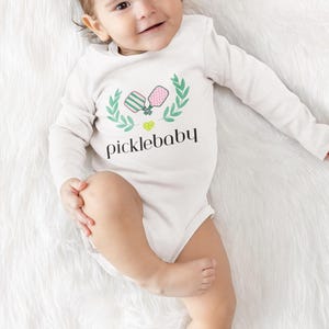 Könnte beinhalten: Ein weißer Baby-Body mit langen Ärmeln und dem Text "picklebaby" in schwarzer Schrift. Über dem Text befindet sich ein grünes und rosa Pickleball-Design mit einem grünen Lorbeerkranz. Das Baby liegt auf einer weißen, flauschigen Oberfläche.
