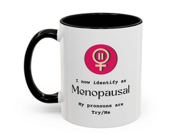 May include: White ceramic mug with a black handle and interior. The mug features a pink and white graphic with a female symbol and the text "I now identify as Menopausal. My pronouns are Try/Me."