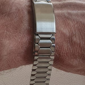 May include: A silver-toned metal watch band featuring a series of interconnected hexagonal links and a smooth, rectangular folding clasp mechanism. The band is constructed from polished stainless steel segments that form a flexible, durable strap.