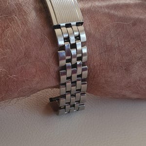 Peut inclure: Un bracelet de montre en métal argenté doté d'un design à maillons multiples et d'un fermoir rectangulaire avec des détails rainurés verticaux. Le bracelet est composé de maillons en acier inoxydable poli avec une boucle déployante sécurisée.