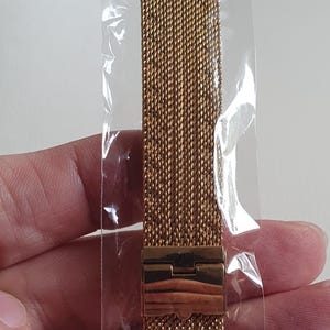 May include: A gold-toned metal watch band featuring a fine mesh weave design and a rectangular folding clasp. The watch strap is encased in a clear plastic sleeve and displays the number 2 in a red font on the upper portion of the band.
