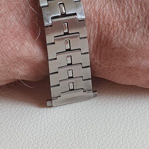 May include: A silver-toned stainless steel watch bracelet featuring a series of interlocking rectangular links and a fold-over clasp mechanism. The metal band has a brushed finish and is designed to connect to a watch case at one end.